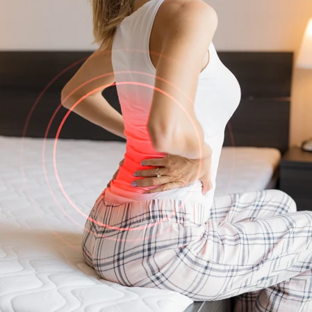 Person holding their back in pain while sitting on a bed.
